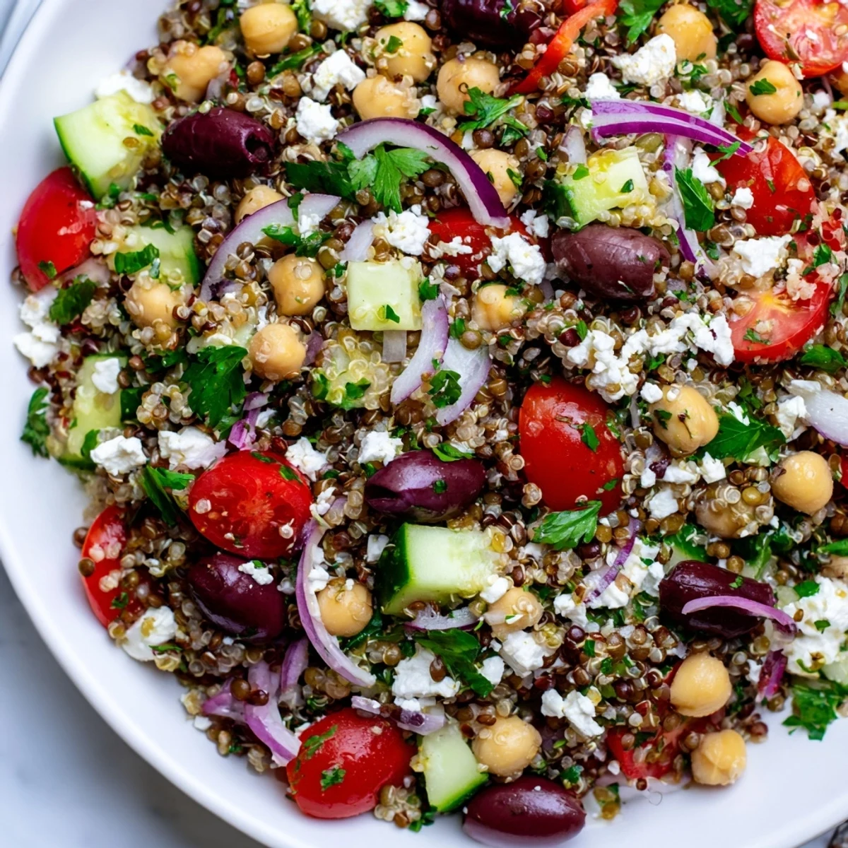 A close-up of Greek Power Salad in a white bowl, showcasing red quinoa, green lentils, chickpeas, and vibrant cherry tomatoes tossed in a zesty vinaigrette.