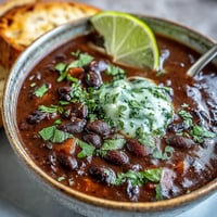 Smoky black bean and sweet potato soup with a zesty lime crema, garnished with fresh cilantro and jalapeño slices.  